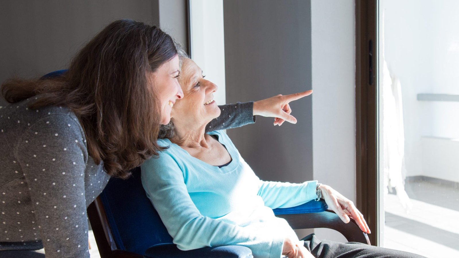 Positive mother and daughter enjoying dramatic view out of window. Young health visitor showing to senior lady funny scene out of window. Family relations concept